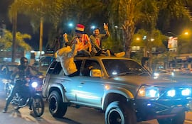 Dos personas con camisetas rojas y blancas celebran en el techo de un vehículo mientras un hombre observa desde una motocicleta.