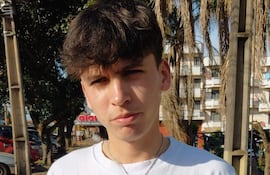 Elías Ríos, joven de cabello oscuro y camiseta blanca, mira a la cámara en una calle, rodeado de árboles y edificios.