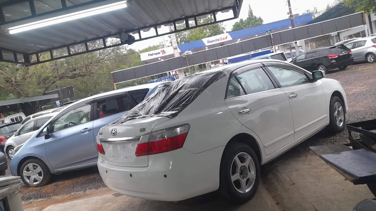 Automóvil sedán blanco con toldo negro en taller, y varios vehículos estacionados con pavimento húmedo.