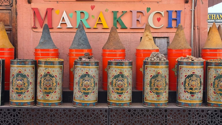 Cilindros cónicos de especias en vibrantes colores, sobre fondo cálido con la palabra 'MARRAKECH' en letras brillantes.