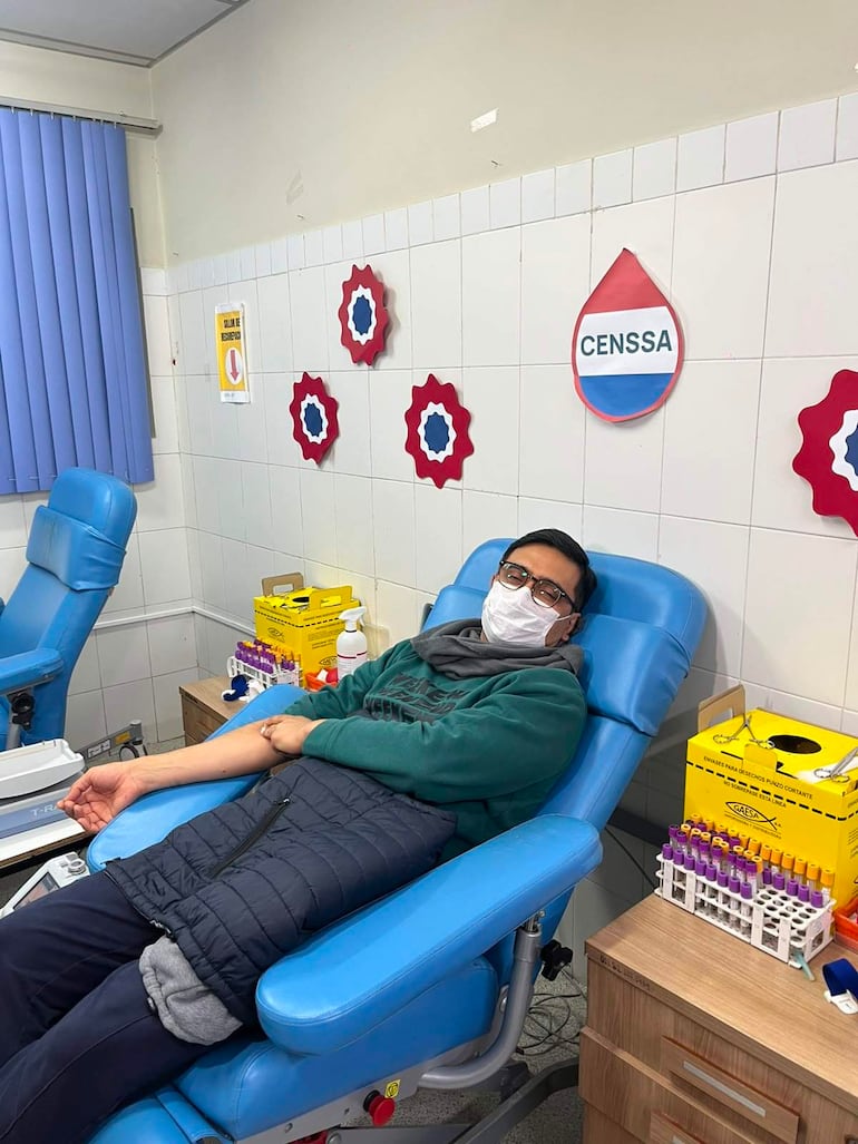 Hombre en suéter verde y chaleco negro reclinado en silla azul, donando sangre con máscara facial en ambiente de salud.