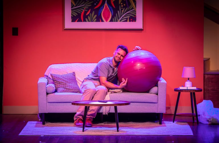 Hombre con cabello corto y camiseta gris abraza una pelota púrpura, sentado en sofá gris con manta blanca, en ambiente interior.