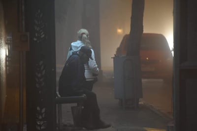 Mujer rubia de pie y hombre sentado en un banco con gorra, ambos rodeados por niebla densa en calle urbana.