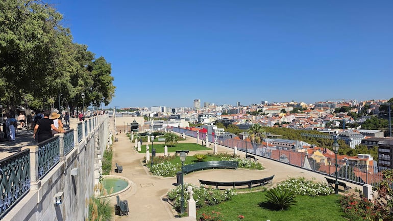 Varias personas pasean y descansan en bancos del parque, rodeados de flores y farolas, con vista a la ciudad.
