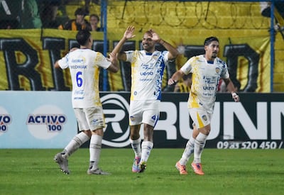 Jugadores de Guarani con camisetas blanco y amarillo celebran con entusiasmo, mientras la multitud vitorea en el fondo.