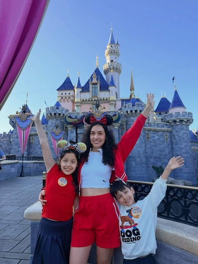 Dos niños con camisetas coloridas y una mujer en atuendo deportivo, sonríen mientras posan frente a un castillo en un día soleado.