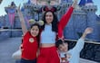 Dos niños con camisetas coloridas y una mujer en atuendo deportivo, sonríen mientras posan frente a un castillo en un día soleado.