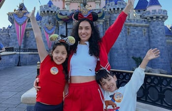 Dos niños con camisetas coloridas y una mujer en atuendo deportivo, sonríen mientras posan frente a un castillo en un día soleado.