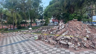 Montículo de escombros en plaza con cinta de advertencia amarilla, rodeado de árboles y edificios de fondo.