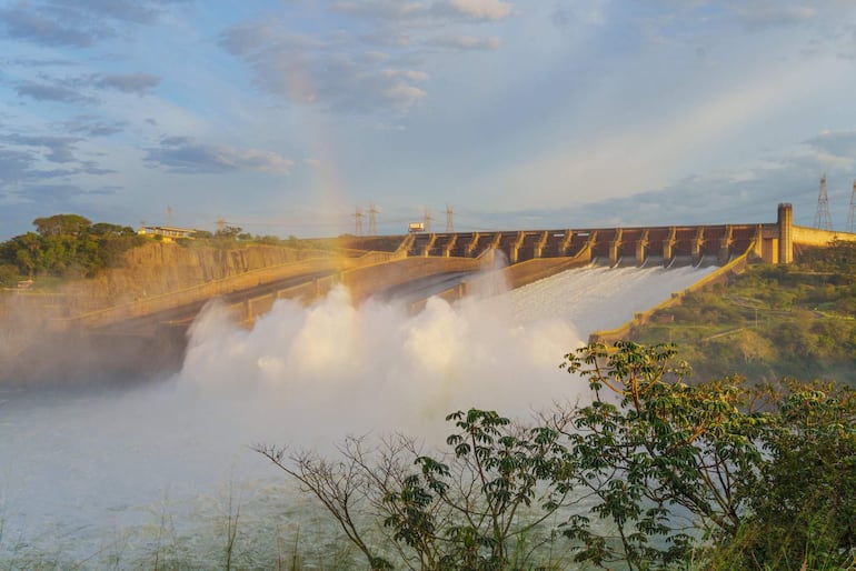 Muro de represa con agua fluyendo violentamente desde compuertas, creando niebla, y torres eléctricas al fondo bajo un cielo nublado.