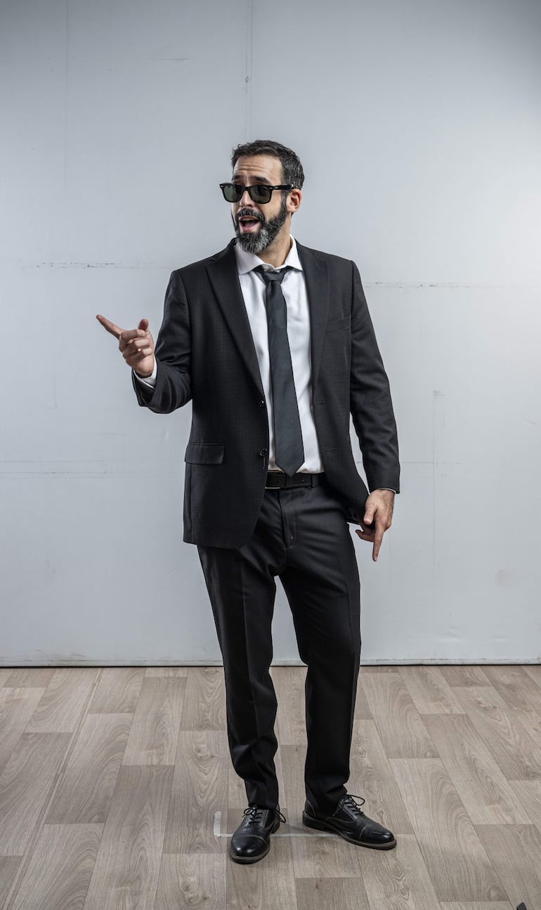 Hombre de pie en traje negro y corbata oscura, señalando con la mano en un fondo gris claro.