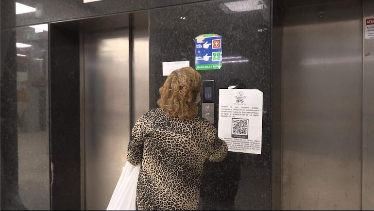 Mujer con cabello rizado y ropa de leopardo sostiene una bolsa blanca frente a panel de ascensores en Hospital Central IPS.