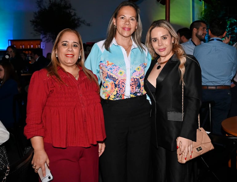 Tres mujeres sonrientes posan en un entorno decorativo, una lleva blusa roja, otra blanca con flores y la última blusa negra con bolso claro.