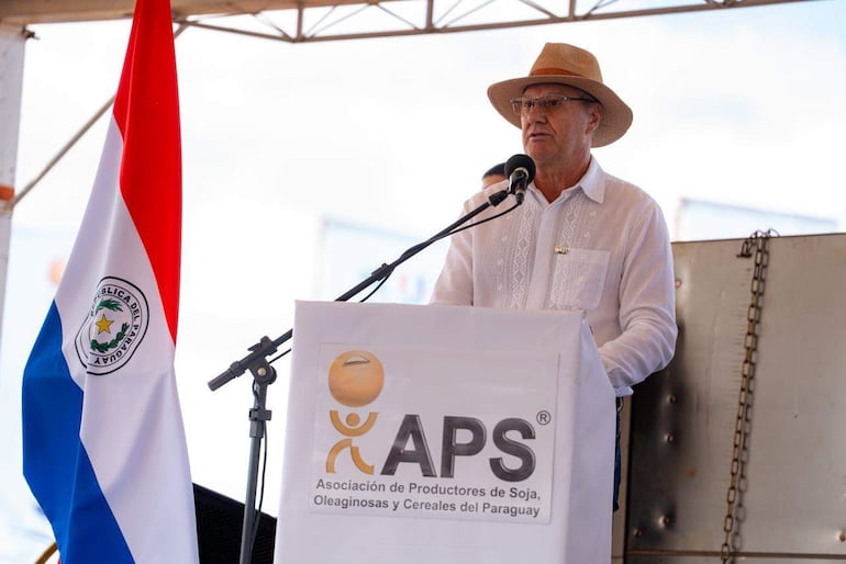 Hombre con sombrero de paja y camisa blanca habla al micrófono, con bandera paraguaya de fondo en un evento agrícola.