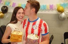 Joshua Duerksen sonríe con camiseta de rayas, mientras Anneke Rahn luce un vestido blanco y sostiene un pastel con velas.