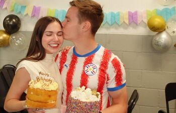 Joshua Duerksen sonríe con camiseta de rayas, mientras Anneke Rahn luce un vestido blanco y sostiene un pastel con velas.