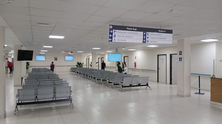Interior del hospital con sillas vacías y una persona vestida de rojo dirigiéndose a una puerta. Espacio bien iluminado y moderno.