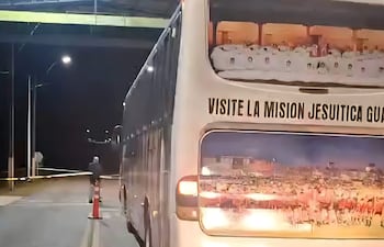 Autobús con cartel que dice 'VISITE LA MISION JESUITICA GUAR' estacionado bajo un puente en un entorno urbano nocturno.