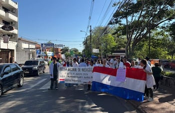 Grupo de médicos en batas blancas sostiene un cartel de huelga y una bandera paraguaya, en una calle urbana con vehículos.