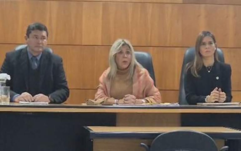 Gloria Hermosa, con blusa marrón y abrigo claro, escucha atentamente junto a Darío Báez y Natalia Cacavelos en un ambiente formal.