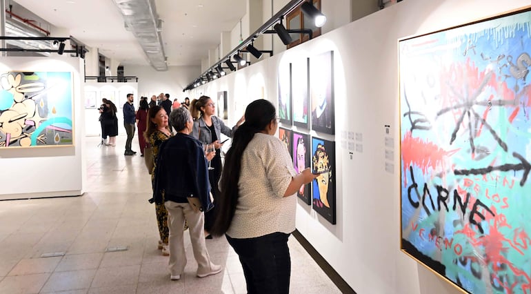 Grupo de personas en galería de arte, algunas conversan, otras observan cuadros abstractos y coloridos en un ambiente iluminado.