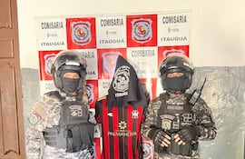 Hombre en camiseta de Club Sportivo Luqueño flanqueado por agentes de policía con cascos, en ambiente de comisaría con carteles de 'COMISARÍA'.