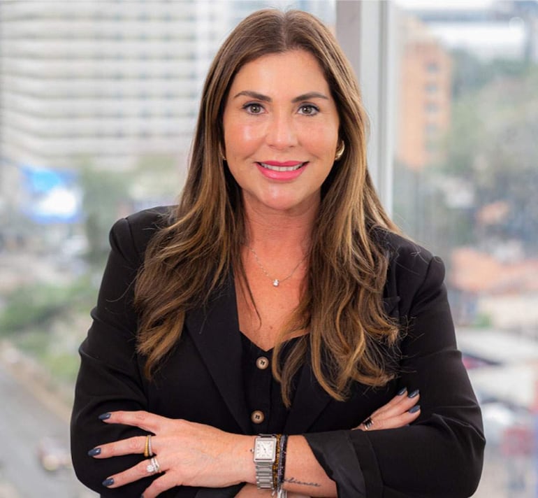 Mujer de cabello rubio y blazer negro sonríe con confianza, de pie frente a un fondo urbano con edificios.