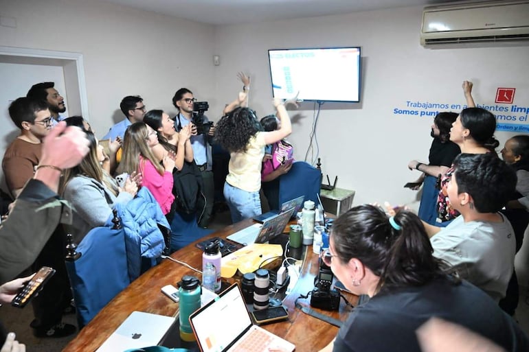 Soledad Núñez levanta los brazos en celebración rodeada de periodistas aplaudiendo en un ambiente festivo.