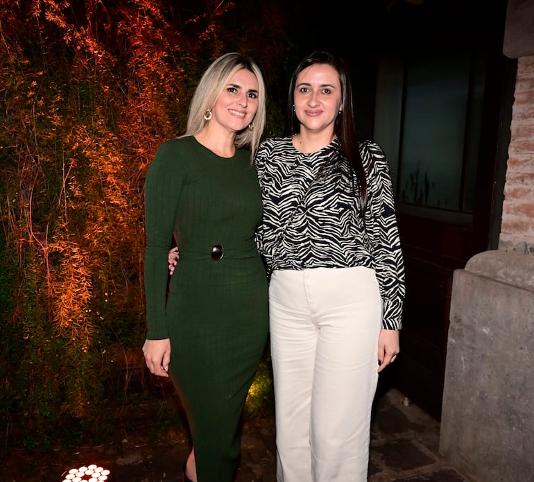 Dos mujeres sonrientes, una con vestido verde y otra con blusa cebra, en un ambiente iluminado con luces cálidas.