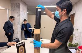 Hombre con guantes y mascarilla inspecciona un objeto en una mesa marcada con 'SENAD'. Dos agentes fiscales y antidrogas observan el operativo.