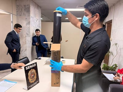 Hombre con guantes y mascarilla inspecciona un objeto en una mesa marcada con 'SENAD'. Dos agentes fiscales y antidrogas observan el operativo.