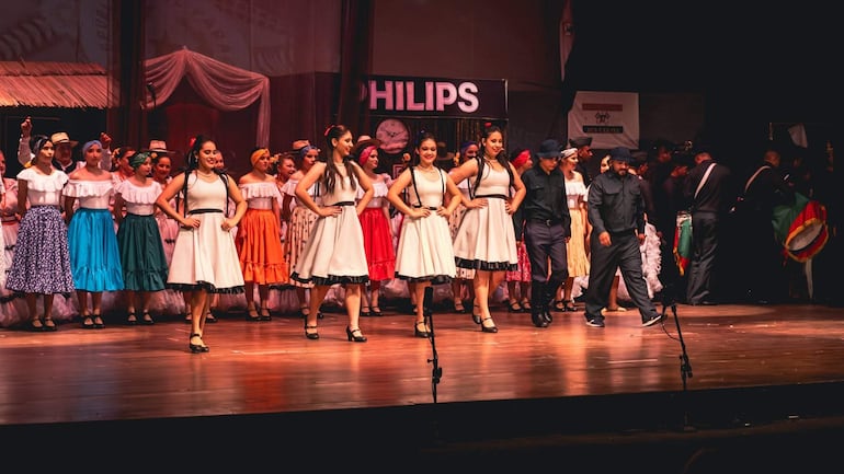 Cinco mujeres con vestidos blancos y negros realizan una danza, mientras otras en trajes coloridos las acompañan en un escenario decorado.