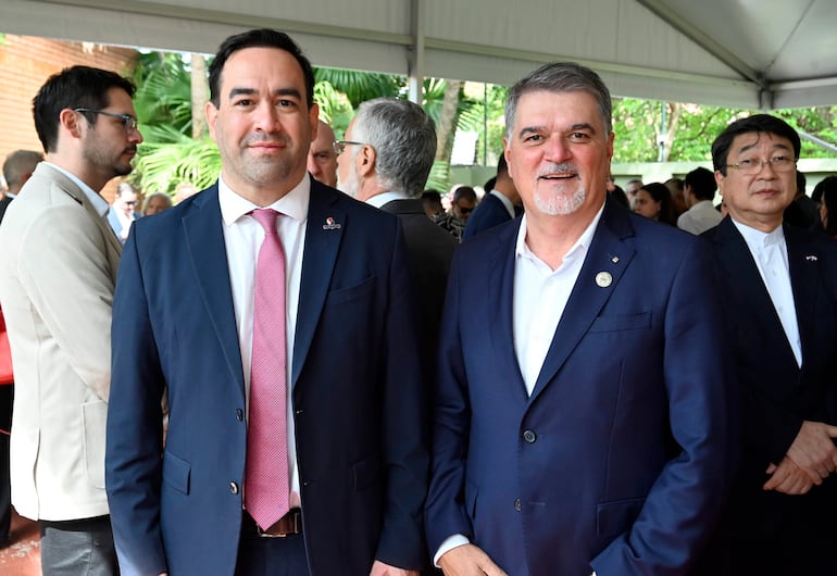 Dos hombres en traje oscuro, uno con corbata rosa y otro con pin, conversan en un evento bajo carpa con fondo de asistentes.
