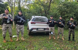 Cuatro hombres armados en uniformes tácticos, listos para una operación, con un vehículo SUV blanco detrás.