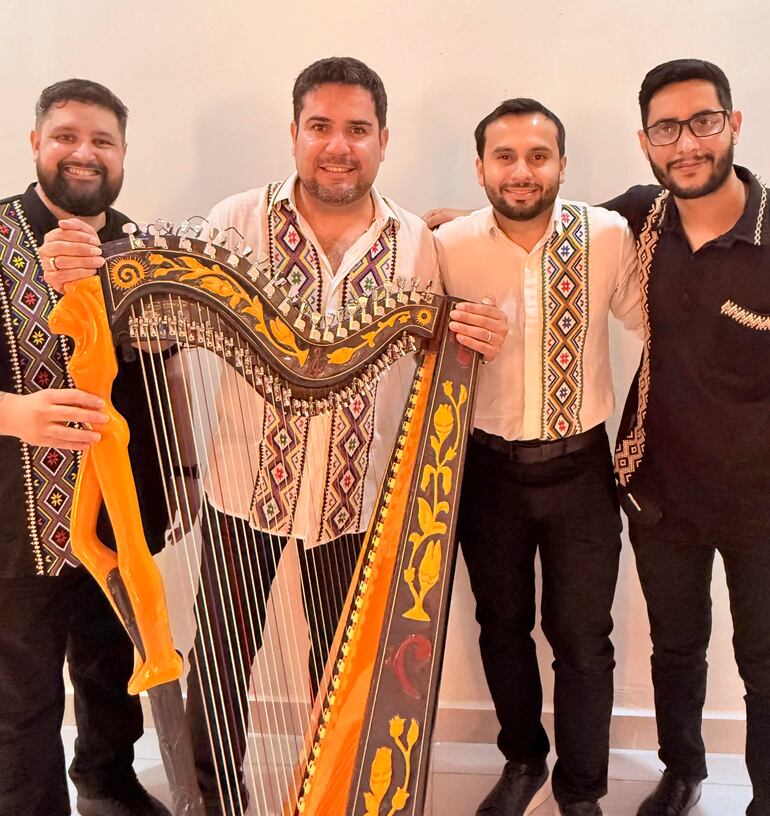 Cuatro hombres sonrientes, dos con camisa negra y dos con camisas blancas, sostienen un arpa en un ambiente claro.