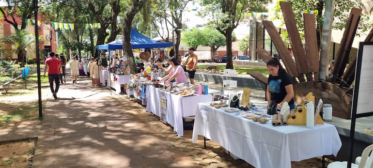 Dos mujeres organizan productos en mesas, mientras un hombre con camiseta roja se dirige a la venta en un soleado entorno de feria.