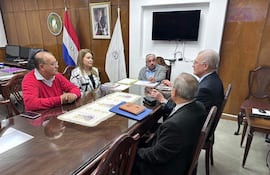 Cinco personas en mesa de oficina: dos hombres en traje, mujer con blusa blanca y negro, discutiendo documentos. Banderas de Paraguay al fondo.