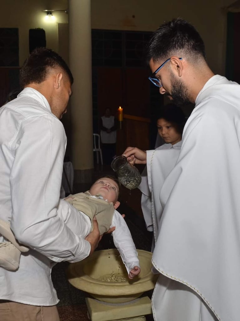 Hombre sostiene a un bebé vestido de blanco, mientras un sacerdote realiza el rito de bautizo en un ambiente religioso sereno.