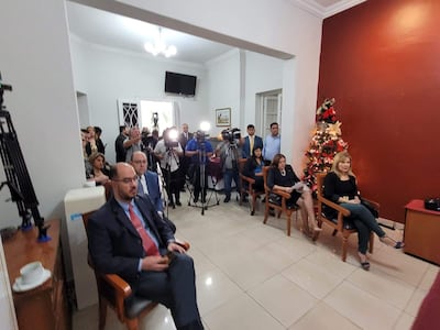 Oscar Paciello en saco oscuro y corbata clara, junto a mujer rubio, rodeados de periodistas y un árbol de Navidad al fondo.