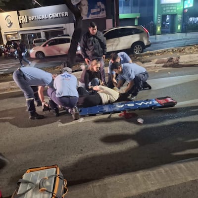 Grupo de seis personas, cuatro en uniformes médicos, atienden a mujer en camilla en la calle frente a un negocio de óptica.