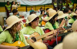 Dos jóvenes, uno tocando guitarra y otro con un teléfono, se preparan para una actuación, vestidos con camisetas verdes y sombreros de paja.