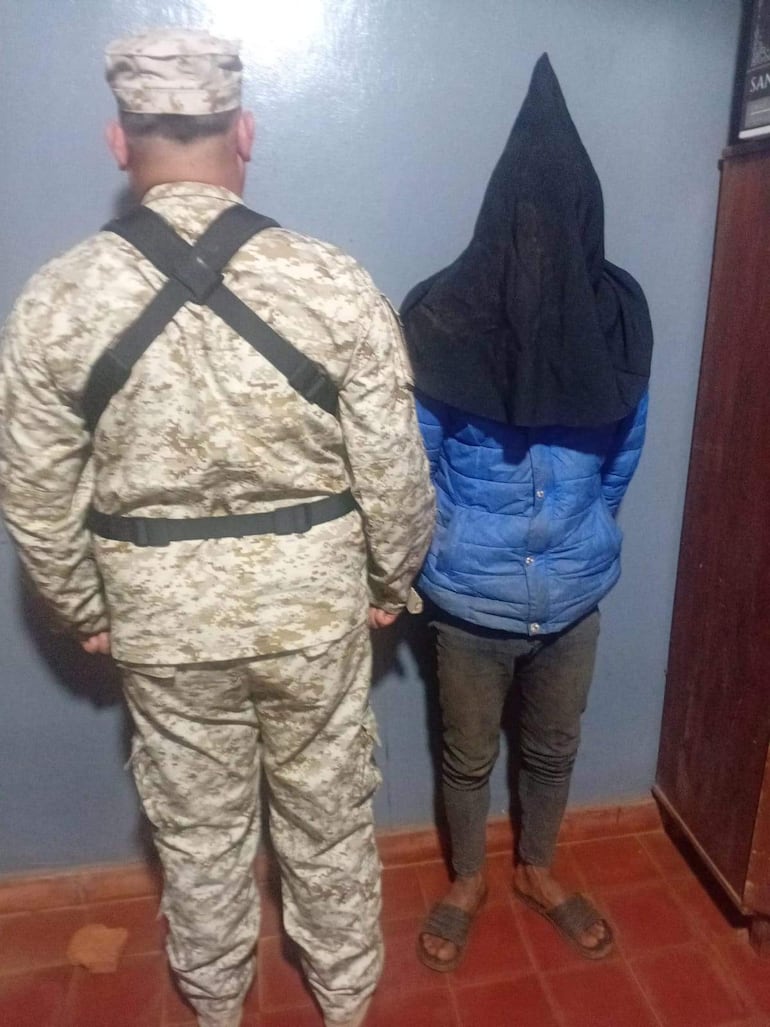Hombre encapuchado a la derecha y policía en fatigues camuflados a la izquierda, en ambiente austero de detención.