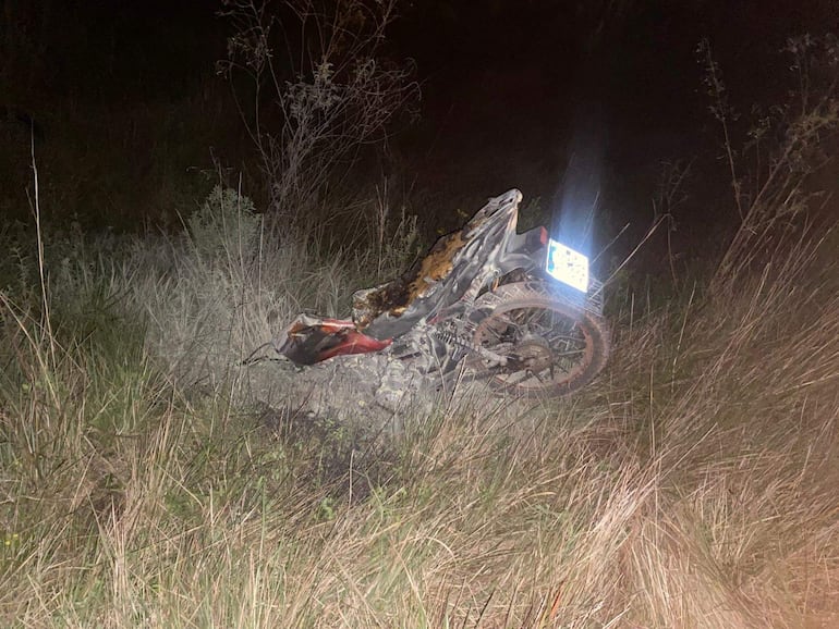 Motocicleta caída en área rural, dañada, rodeada de vegetación y hierba, sin personas visibles.