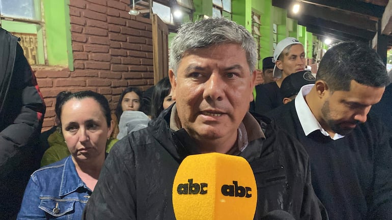 Hombre canoso habla en micrófono, mujer con chaqueta de mezclilla escucha, joven con lluvia de plástico detrás, ambiente exterior.