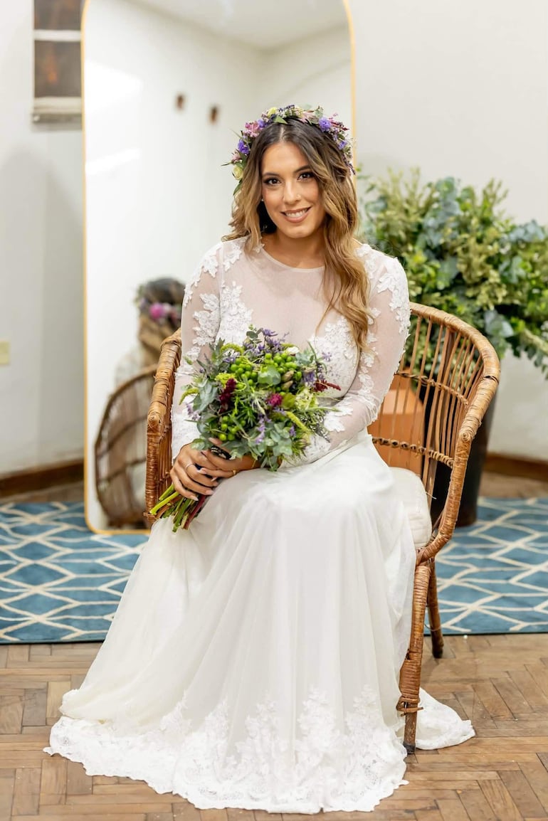 Mujer en vestido blanco con encaje y diadema de flores, sonriendo y sosteniendo un ramo en un entorno acogedor.