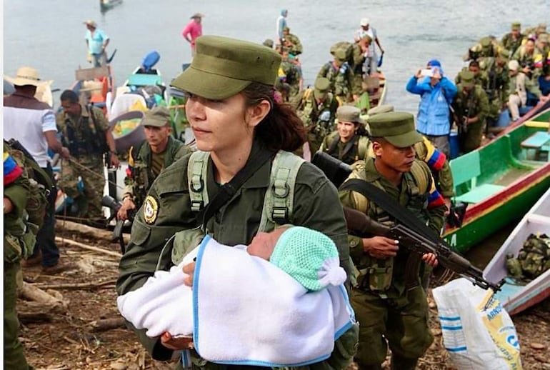 Mujer militar con expresión seria sostiene un bebé en una manta, rodeada de soldados en uniformes, con ambiente de actividad.