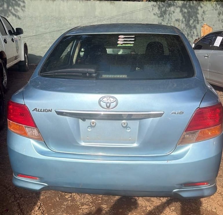 Automóvil Toyota Allion azul estacionado frente a un patrullero en un entorno de tierra y pared gris.