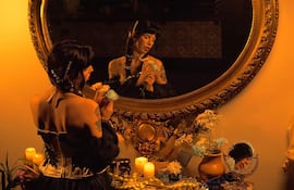 Mujer de espaldas con peinado oscuro y flores en mano, frente a un espejo ornamentado rodeada de velas y objetos decorativos.