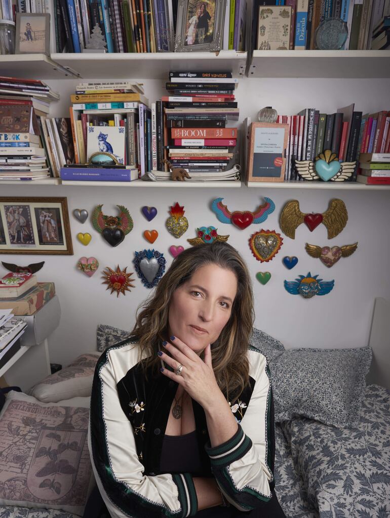 Agustina Bazterrica sentada en una cama con chaqueta negra y expresión reflexiva, rodeada de libros en un ambiente acogedor.