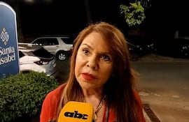 Mujer de cabello largo y suelto, vestida con blusa roja y azul, sostiene un micrófono mientras habla en un ambiente urbano.